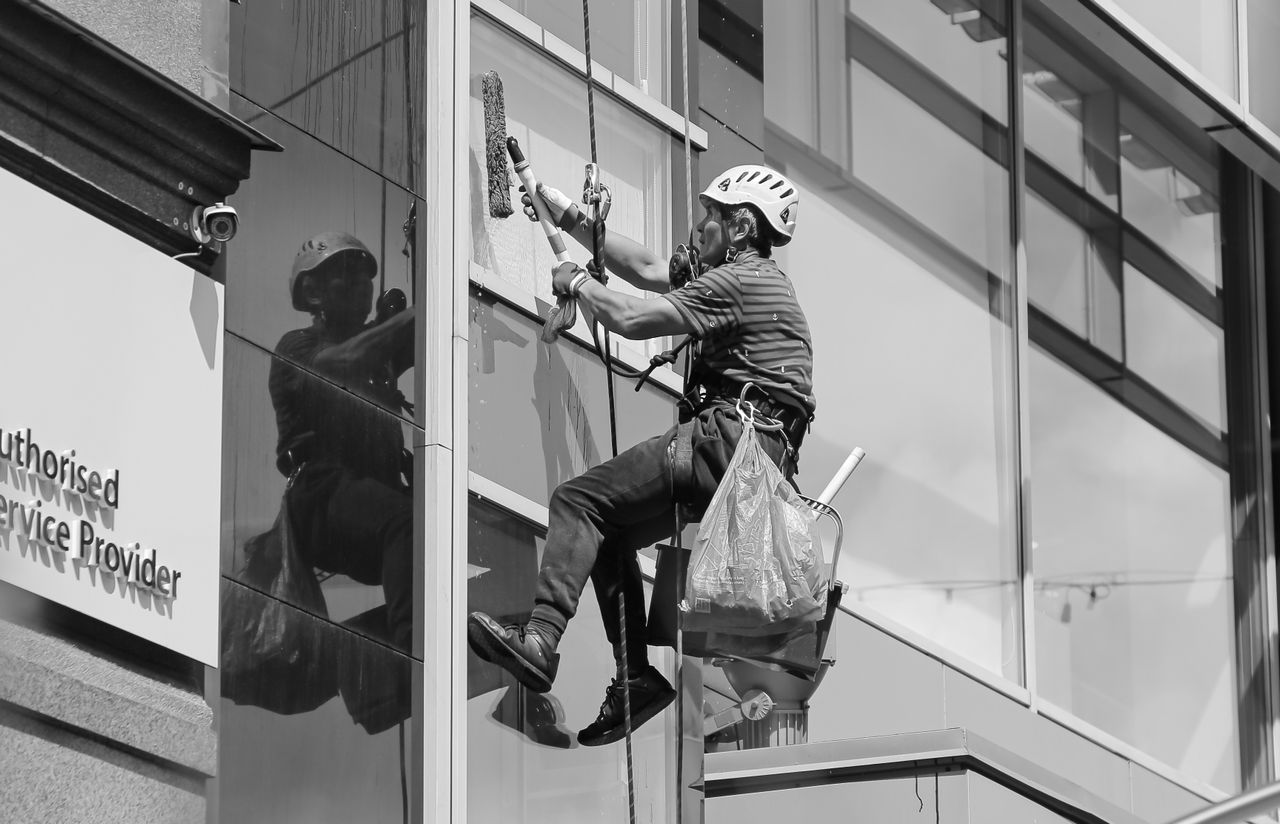 services-04 Black and white from below of man steeplejack in protective helmet with professional equipment cleaning glass wall of modern tower